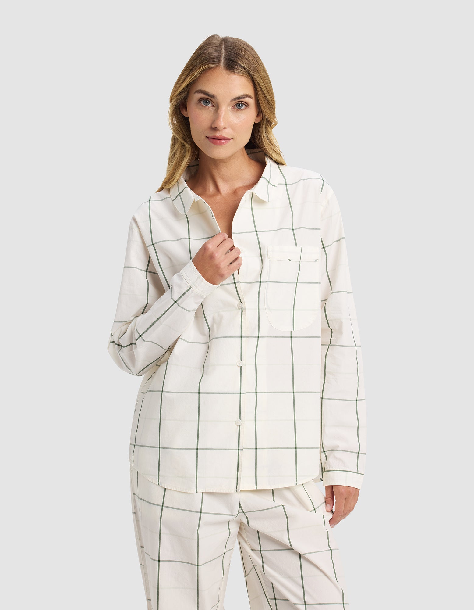 A woman with light brown hair stands against a plain background wearing the Cozy Earth Women's Sutton Long Sleeve Pajama Top in cream with a green windowpane grid pattern, touching her collar with a neutral expression.