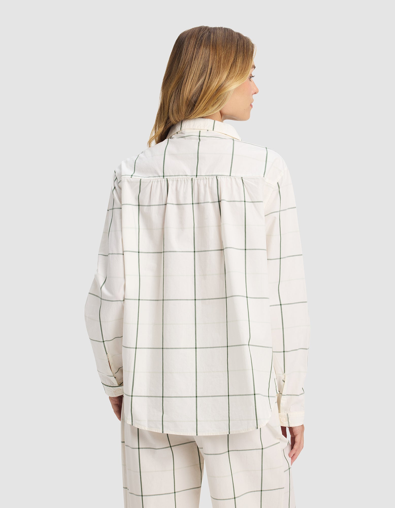 A woman with light brown hair wears the Cozy Earth Women's Sutton Long Sleeve Pajama Top, a loose-fitting white shirt with green grid pattern, collar, and back yoke. She faces away against a plain gray background.