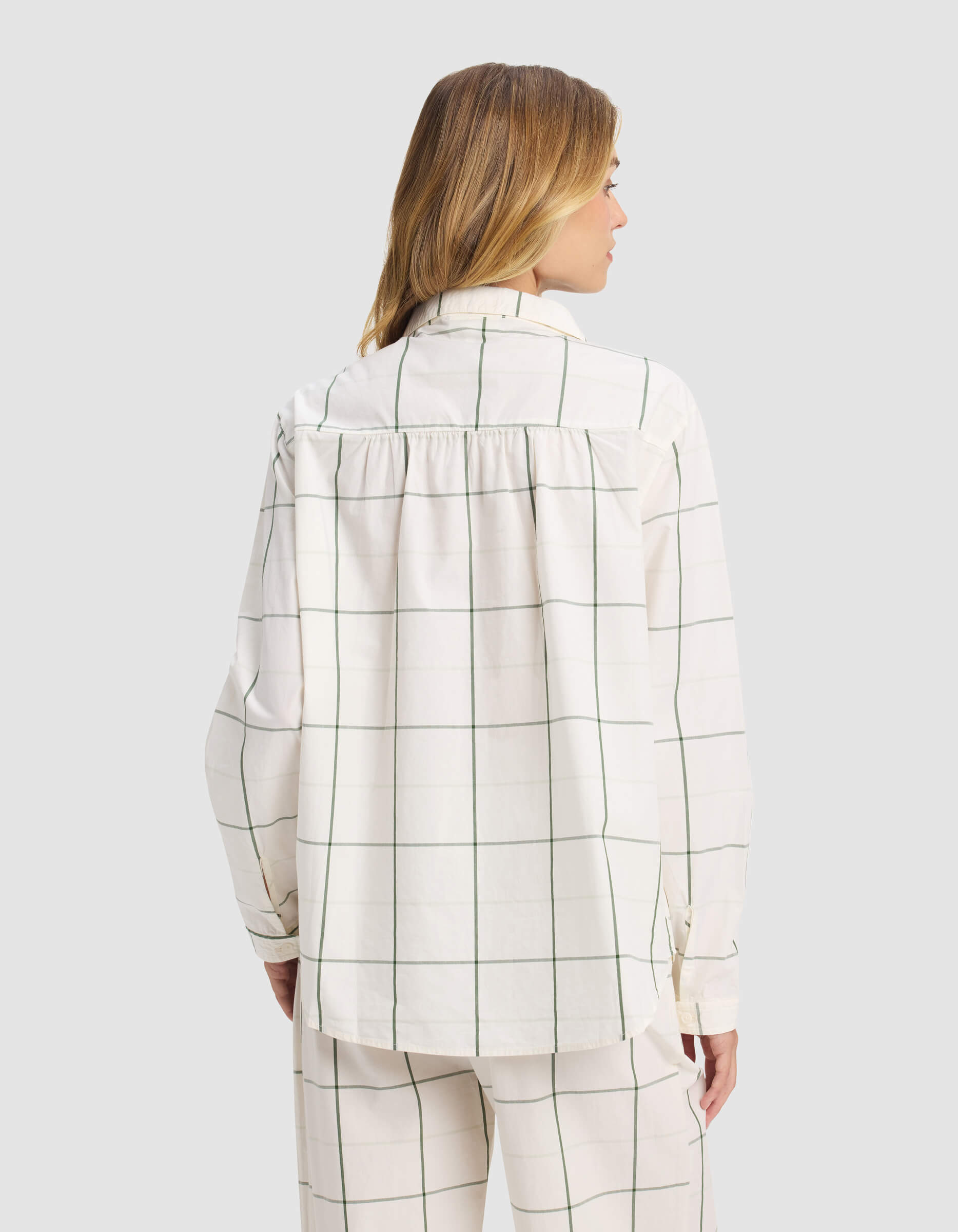 A woman with light brown hair faces away from the camera, wearing the Cozy Earth Women's Sutton Long Sleeve Pajama Top—a loose-fitting, white shirt with a green windowpane grid pattern—along with matching pants.