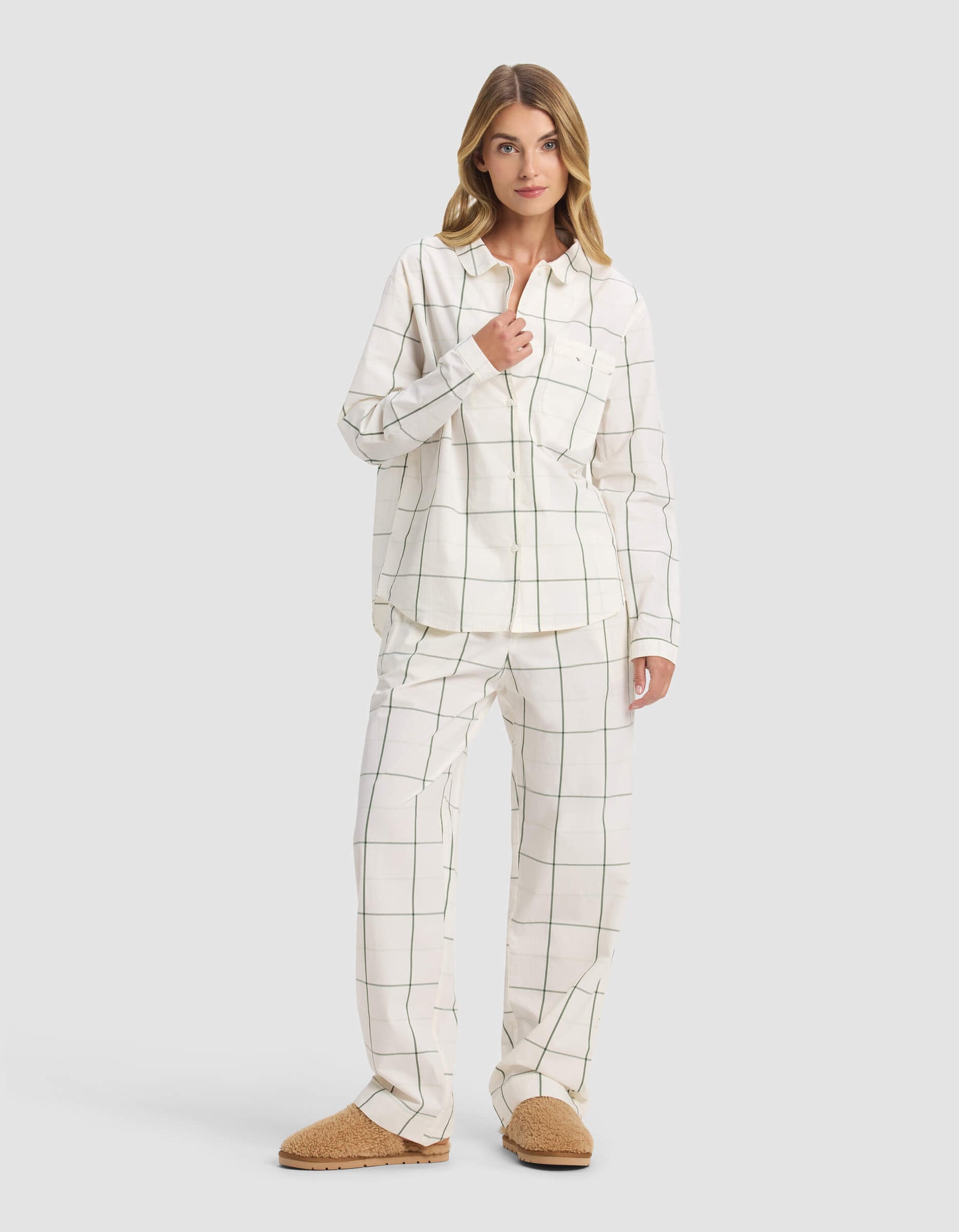 A woman with blonde hair poses casually against a plain background, wearing Cozy Earth's Women's Sutton Long Sleeve Pajama Top in a white and black windowpane check print, paired with matching bottoms and tan slippers.