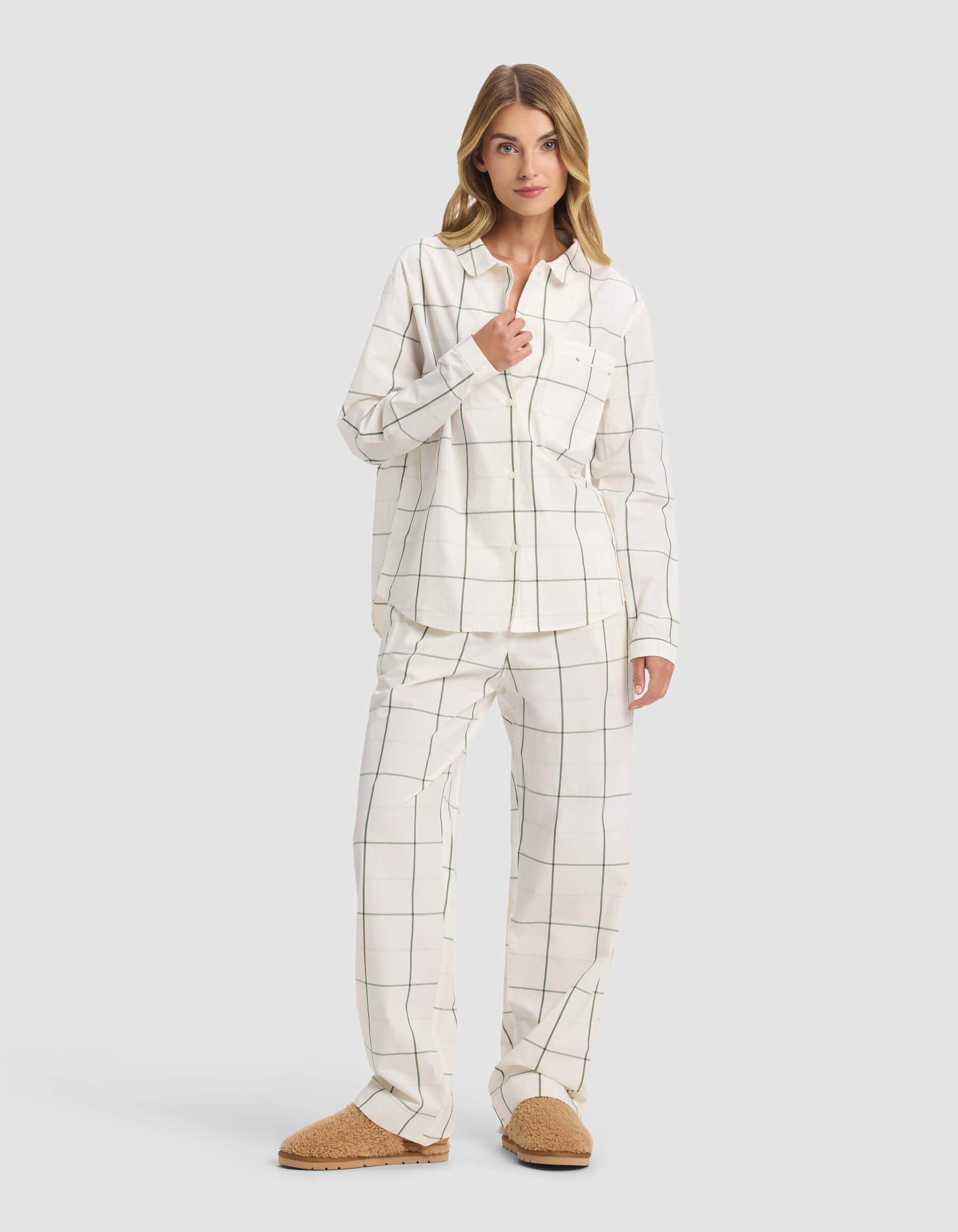 A woman with blonde hair poses casually against a plain background, wearing Cozy Earth's Women's Sutton Long Sleeve Pajama Top in a white and black windowpane check print, paired with matching bottoms and tan slippers.|Color:Bone Windowpane