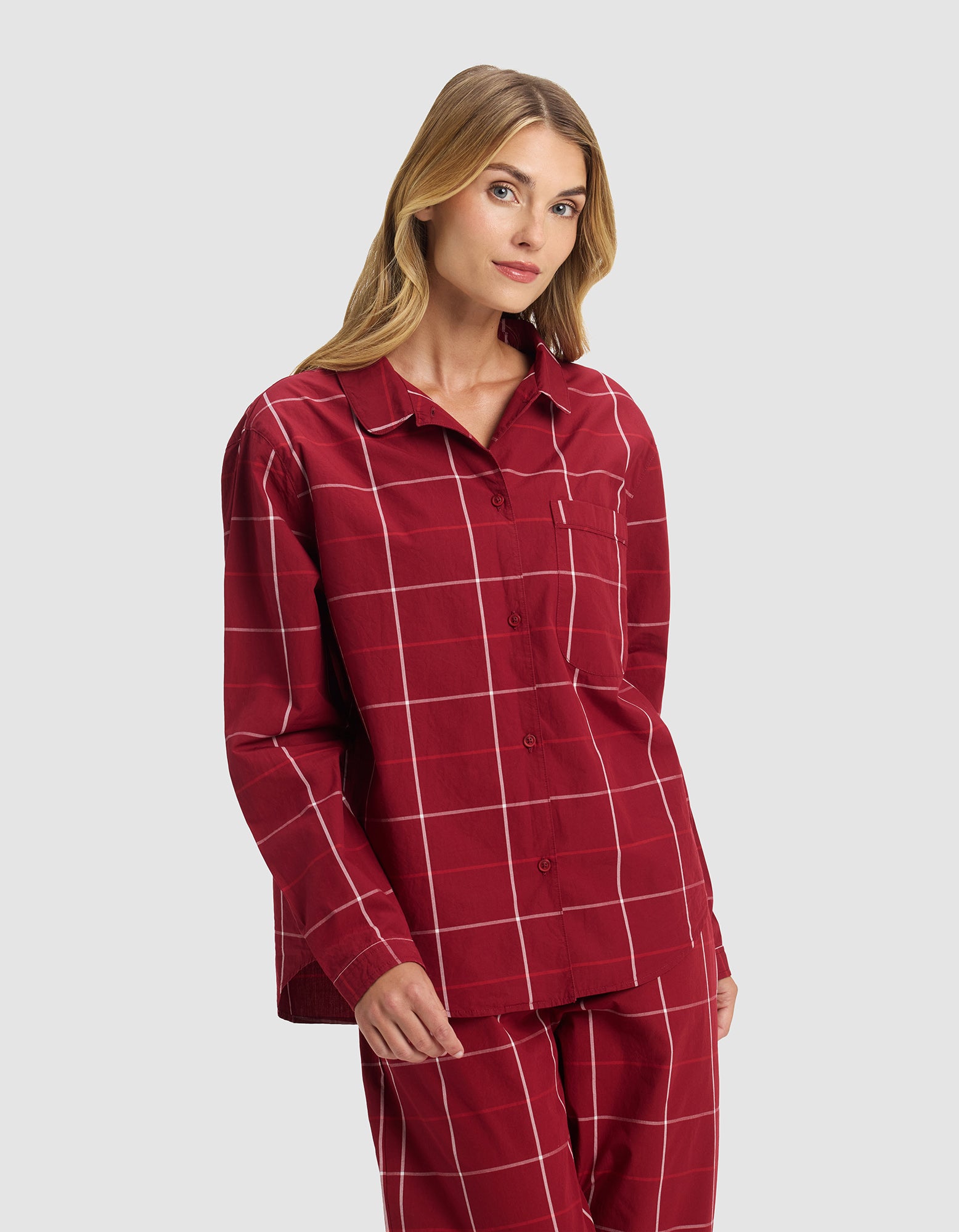 A woman wearing the Cozy Earth Women's Sutton Long Sleeve Pajama Top in red plaid with white grid lines stands against a plain light grey background, looking slightly to the side.|Color:Garnet Windowpane