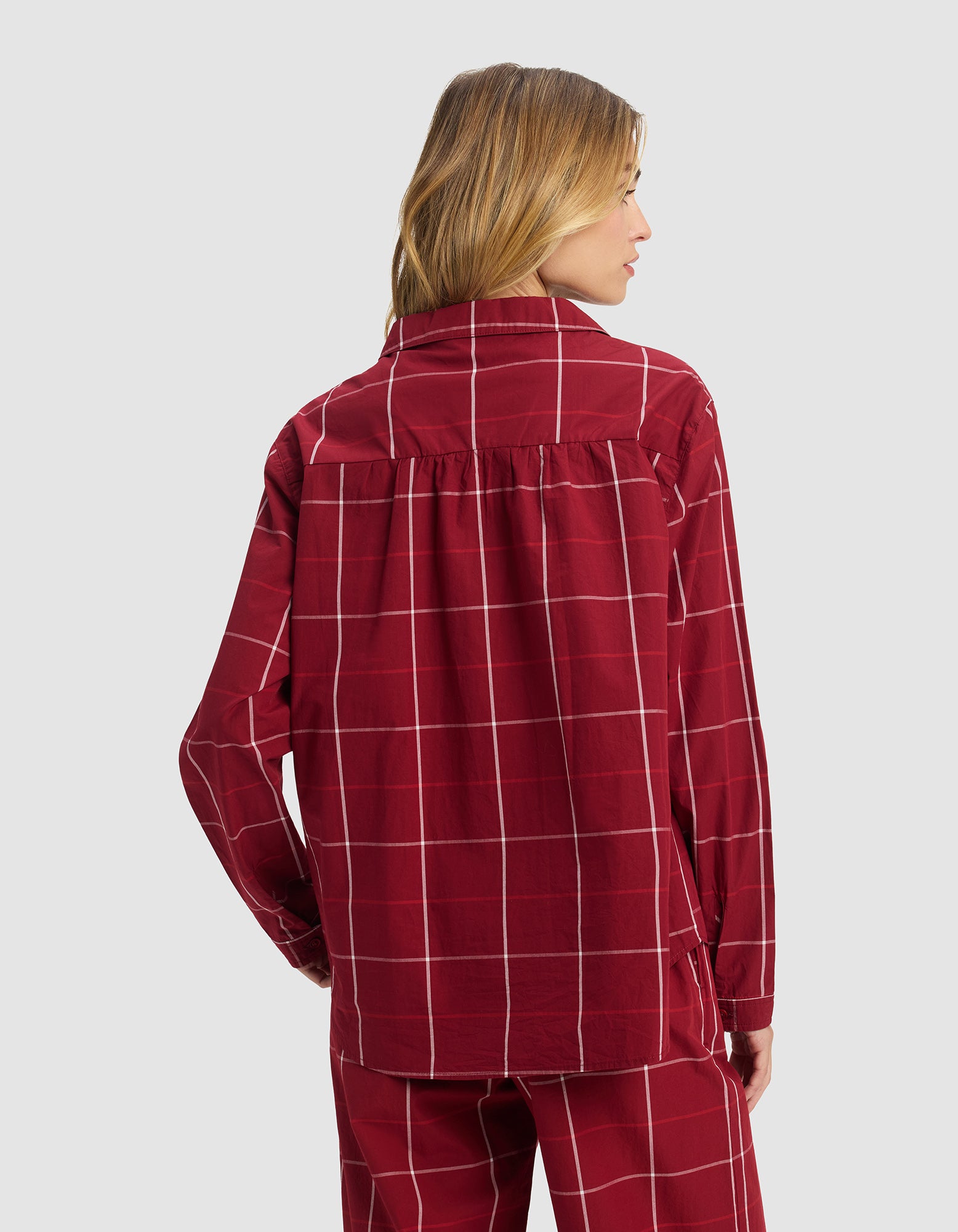 A woman with light brown hair faces away, wearing Cozy Earth's Women's Sutton Long Sleeve Pajama Top in red with a white grid pattern and matching pants, set against a plain light gray background.|Color:Garnet Windowpane