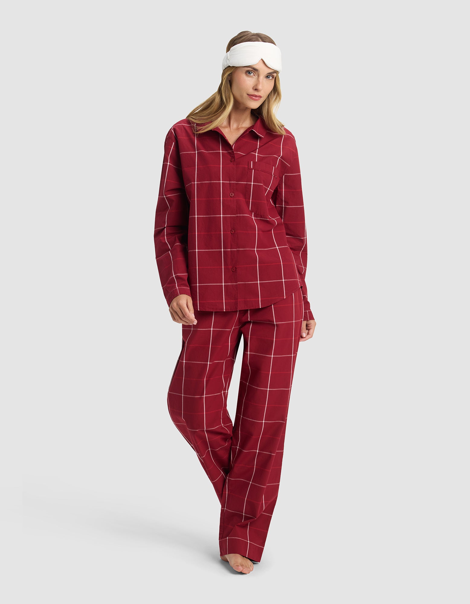 A woman stands barefoot against a light gray background, wearing Cozy Earth's Women's Sutton Long Sleeve Pajama Top in red plaid and a white sleep mask.|Color:Garnet Windowpane