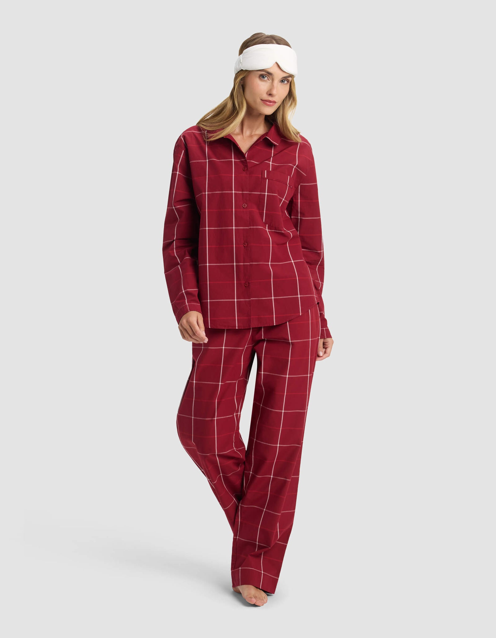 A woman models the Cozy Earth Women's Sutton Long Sleeve Pajama Top, shown in red plaid, paired with a white sleep mask, standing barefoot against a plain gray background.