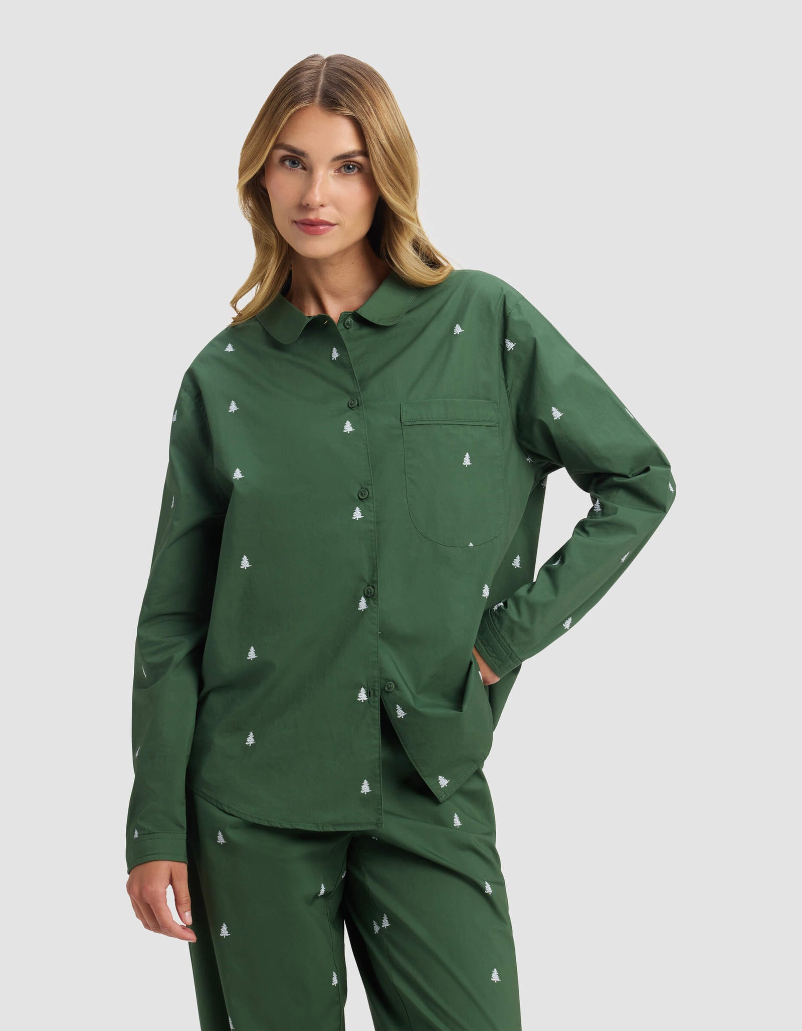 A woman models the Cozy Earth Women's Sutton Long Sleeve Pajama Top, featuring a matching green set with a small white tree pattern, as she poses with one hand in her pocket against a light gray background.