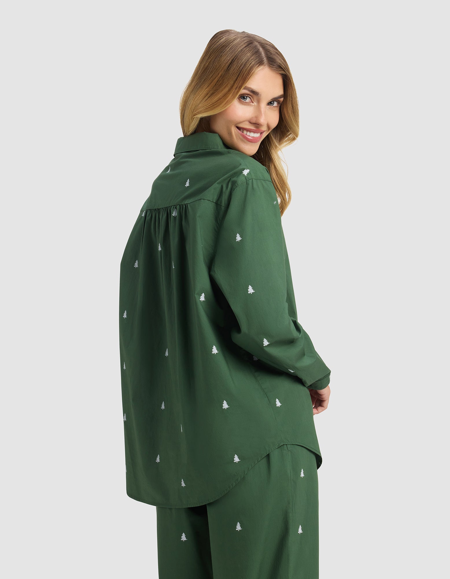 A woman with long blonde hair smiles over her shoulder, wearing the Cozy Earth Women's Sutton Long Sleeve Pajama Top in dark green with a small white tree pattern, paired with matching pants, against a plain light gray background.