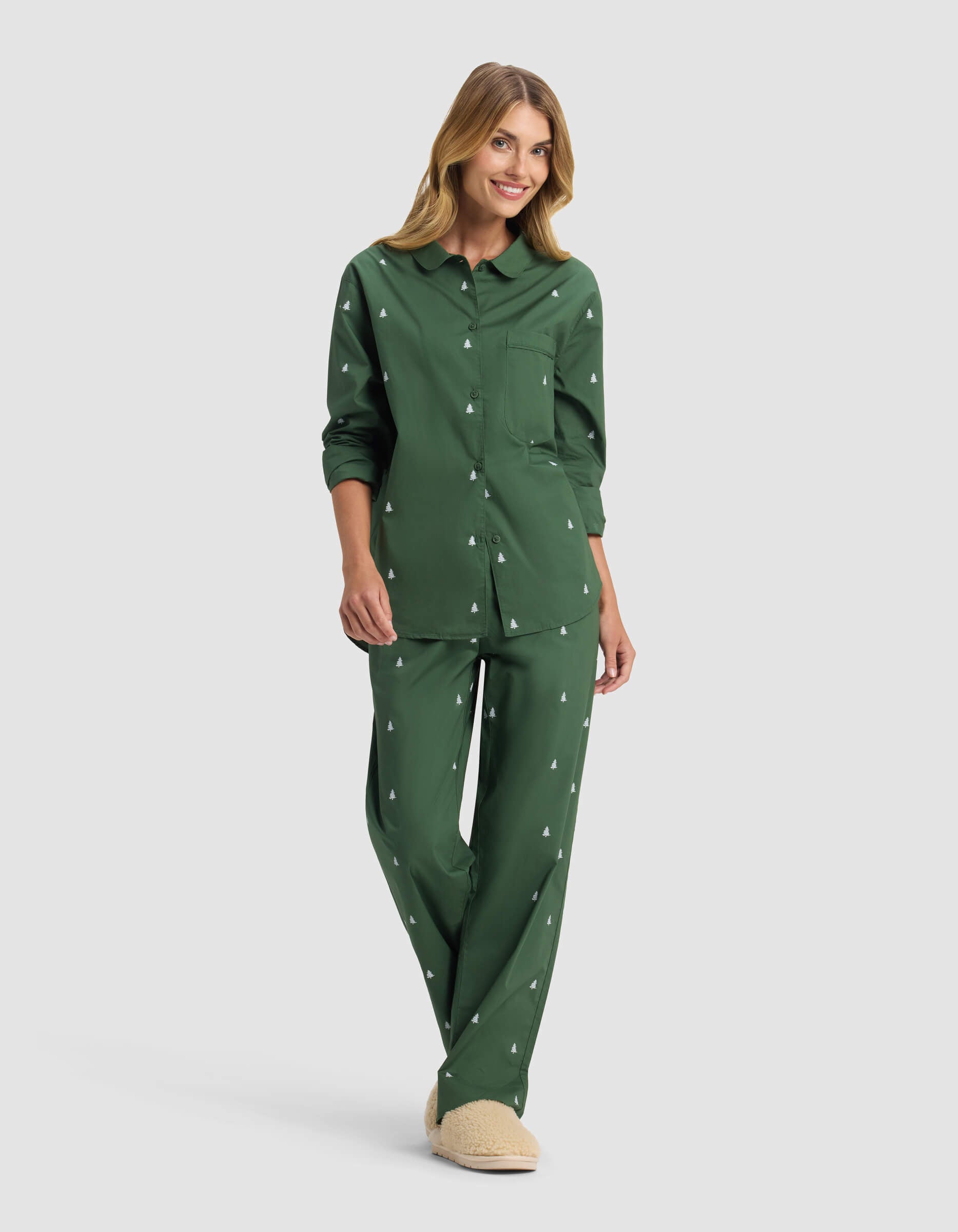 A woman smiles in Cozy Earth's Women's Sutton Long Sleeve Pajama Top, featuring a dark green color with a white tree print, paired with beige slippers, standing against a light gray background.
