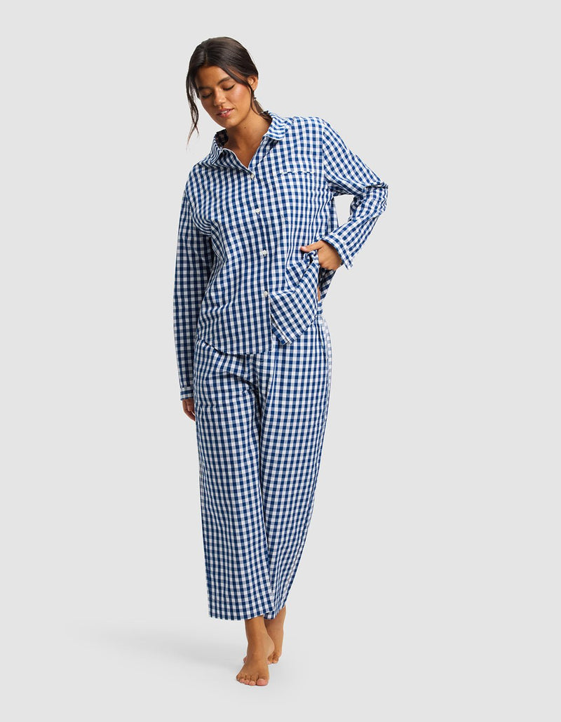 A woman stands barefoot against a plain light background, wearing the Cozy Earth Women's Sutton Long Sleeve Pajama Set, featuring a blue and white checkered button-up top and matching loose-fitting pants.