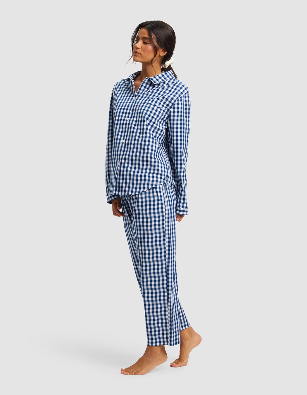 A woman poses barefoot against a light gray background wearing Cozy Earth's Women's Sutton Long Sleeve Pajama Top in blue and white checkered pattern, paired with matching pajama pants.|Color:Pacific Gingham