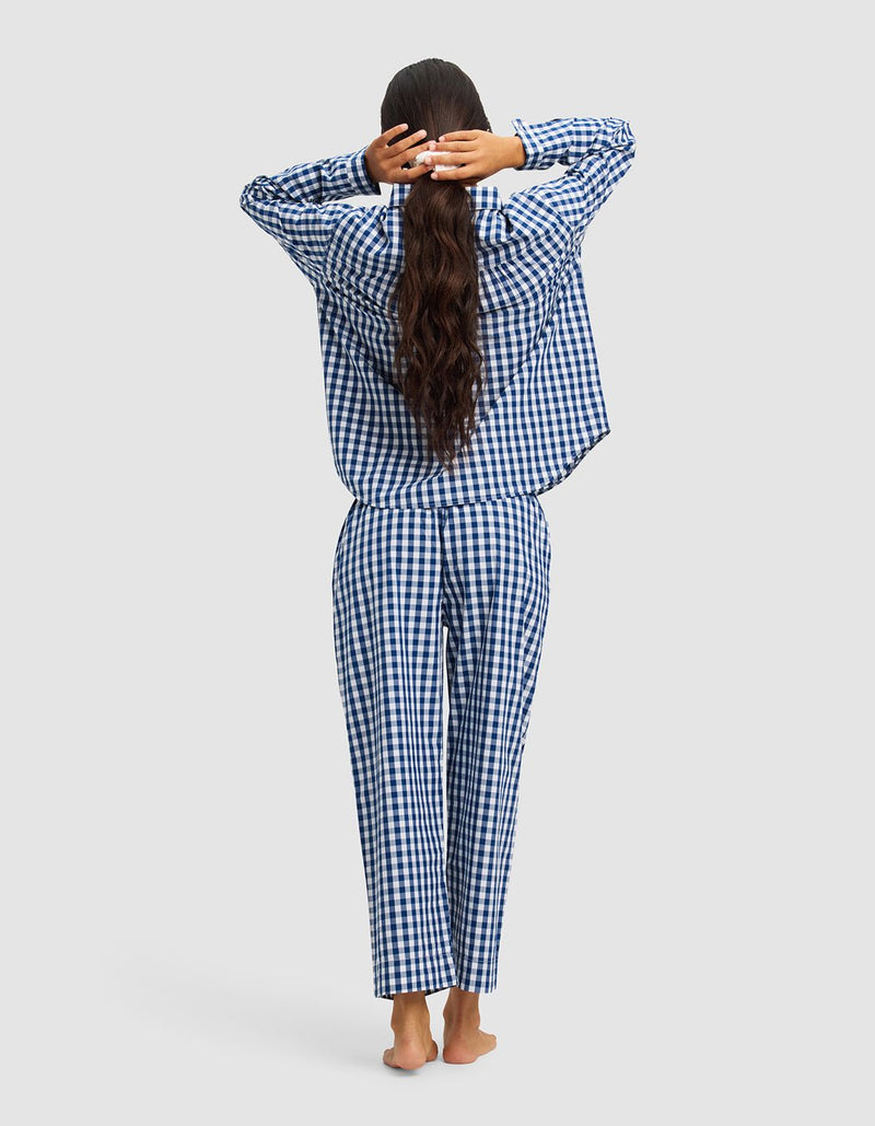 A woman with long, wavy hair stands barefoot facing away, stretching with her hands behind her head while wearing the Cozy Earth Women's Sutton Long Sleeve Pajama Set in blue and white checkered pattern against a light gray background.