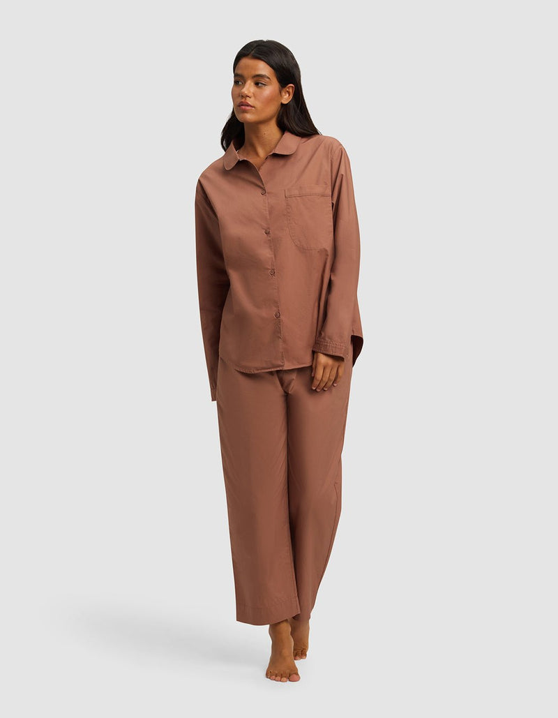 A woman stands barefoot against a plain background, wearing the Cozy Earth Women's Sutton Long Sleeve Pajama Set—a loose-fitting brown button-up top with a chest pocket and matching wide-leg pants.