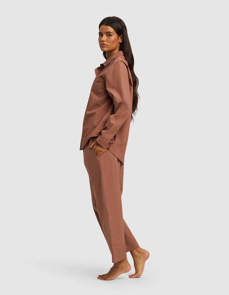 A woman with long dark hair stands barefoot in profile, wearing the Cozy Earth Women's Sutton Long Sleeve Pajama Set in light brown, her hands in her pockets against a plain light background.
