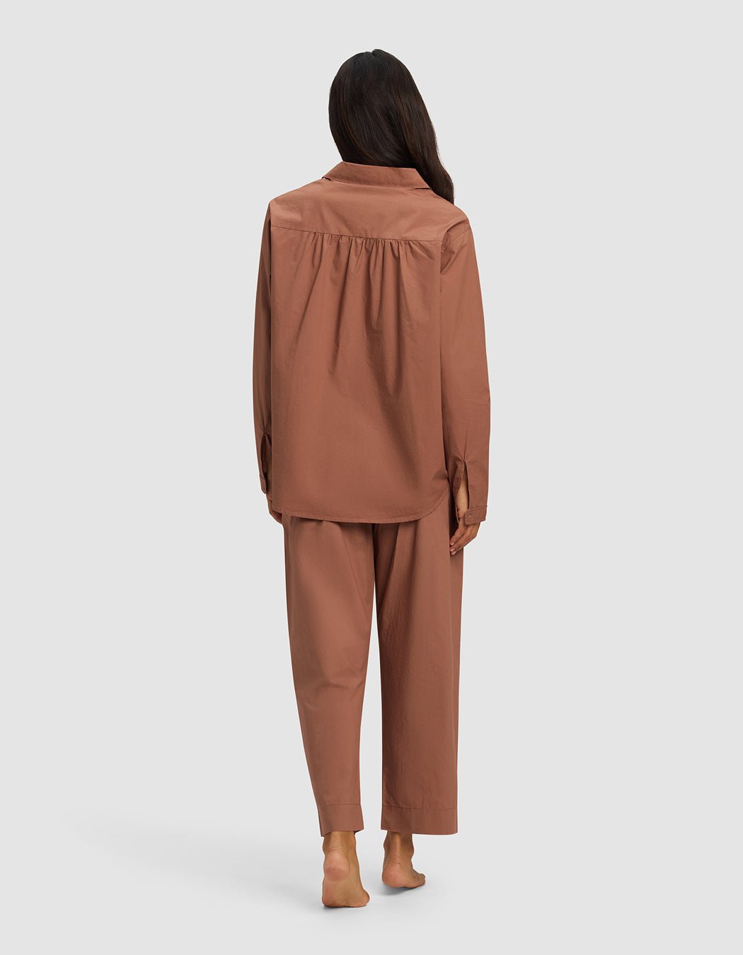 A barefoot woman with long dark hair is shown from behind wearing the Cozy Earth Women's Sutton Long Sleeve Pajama Set in brown, featuring a loose fit and long sleeves and pants, against a plain light gray background.