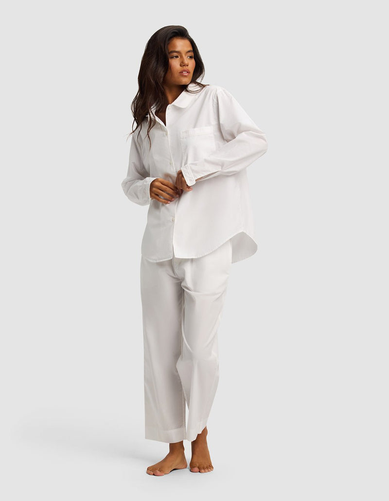 A woman with long dark hair stands barefoot against a light gray background, wearing the Cozy Earth Women's Sutton Long Sleeve Pajama Set—loose-fitting white pajamas featuring a button-up shirt and wide-leg pants.