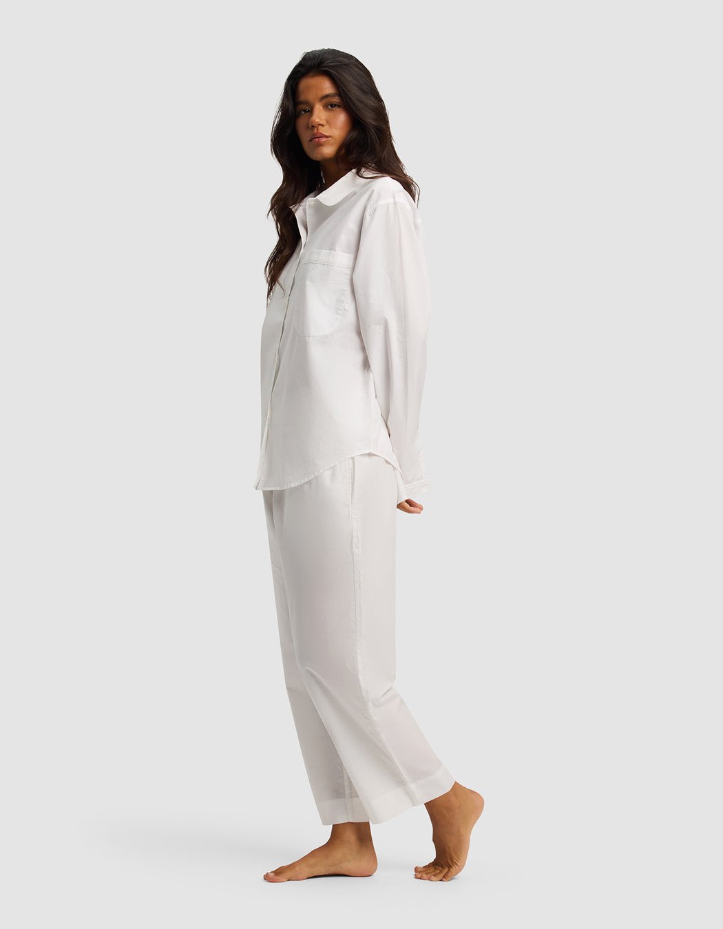 A woman with long dark hair stands barefoot in the Women's Sutton Long Sleeve Pajama Set by Cozy Earth—loose-fitting white shirt and matching pants—against a plain light gray background.