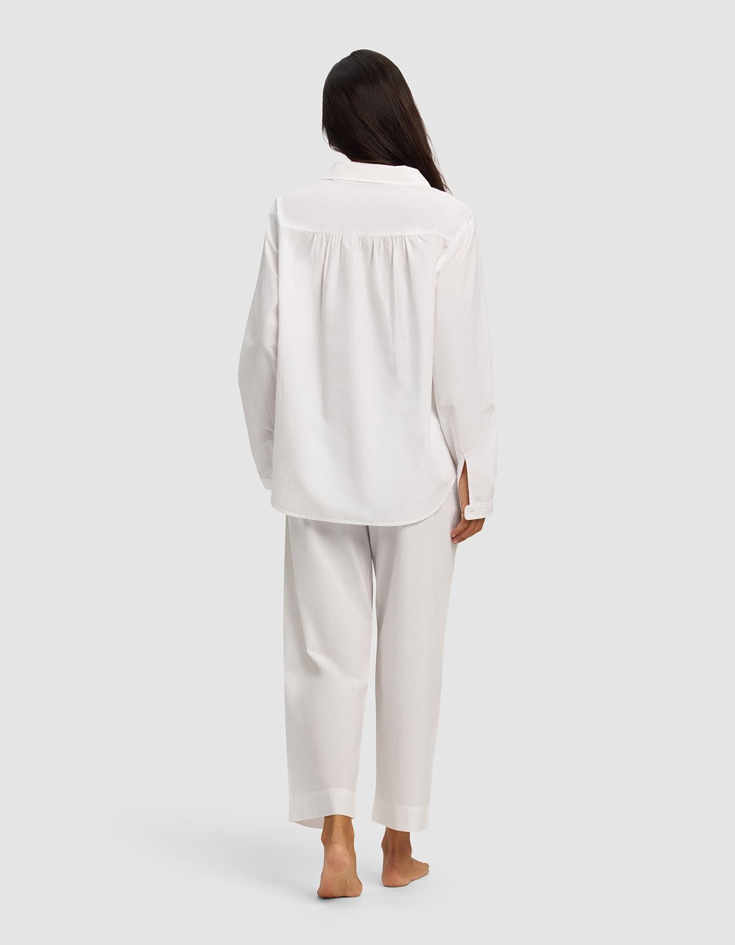 A woman with long dark hair, seen from behind, stands barefoot on a light gray background wearing the Cozy Earth Women's Sutton Long Sleeve Pajama Set in white.
