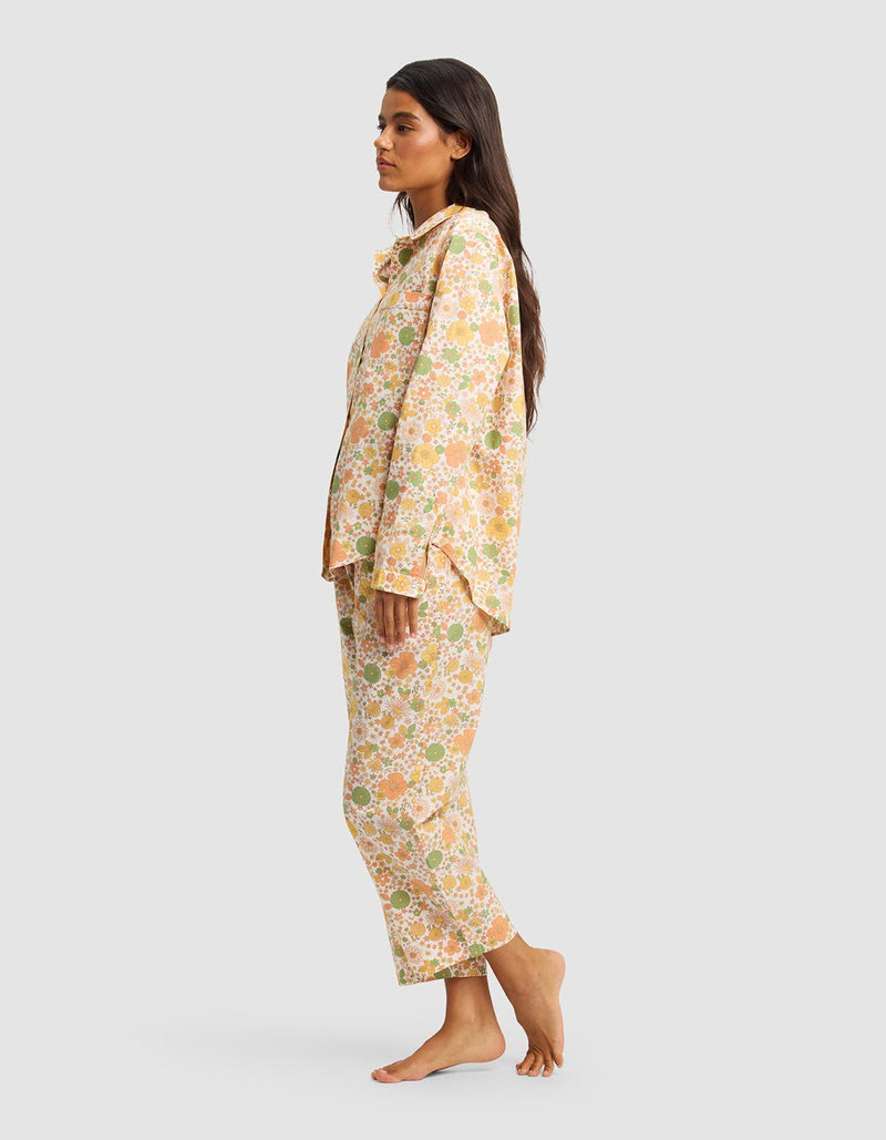 A woman stands sideways barefoot against a plain light gray background, wearing the Cozy Earth Women's Sutton Long Sleeve Pajama Set—loose-fitting, floral-patterned pajamas in shades of peach, yellow, and green.
