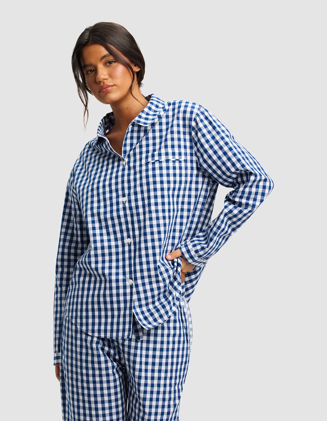 A woman models the Cozy Earth Women's Sutton Long Sleeve Pajama Top, featuring blue and white checks and matching pants, against a plain light gray background.|Color:Pacific Gingham