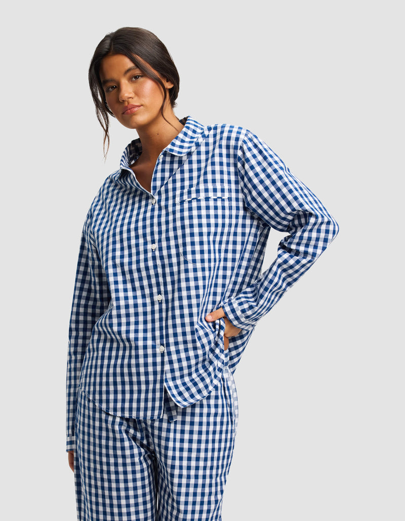 A woman with dark hair models the Women's Sutton Long Sleeve Pajama Top by Cozy Earth in blue and white gingham, standing against a light background with one hand on her hip.