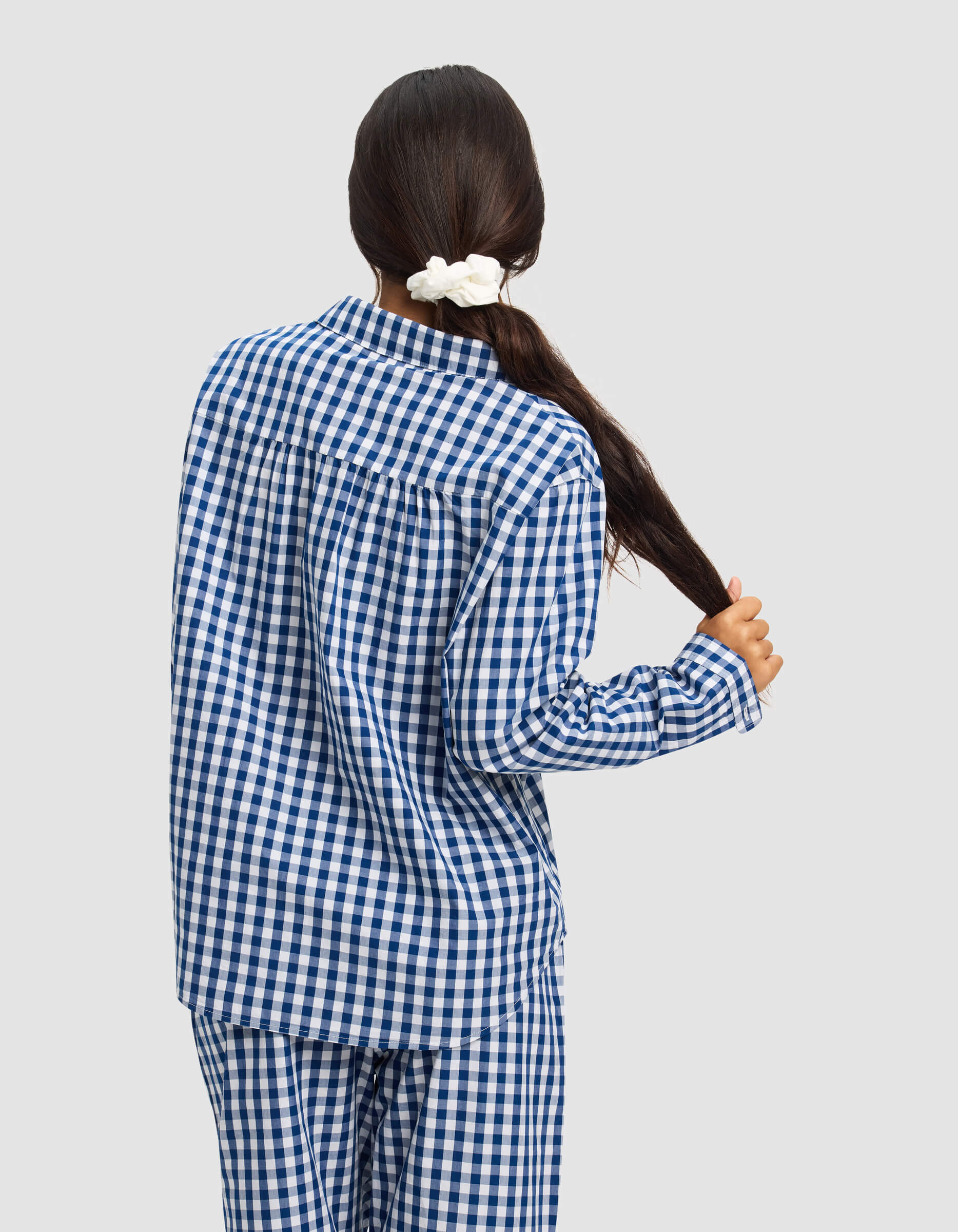 A woman with long dark hair in a white scrunchie, seen from behind holding her braid, wears the Cozy Earth Women's Sutton Long Sleeve Pajama Top in blue and white checks against a plain light background.