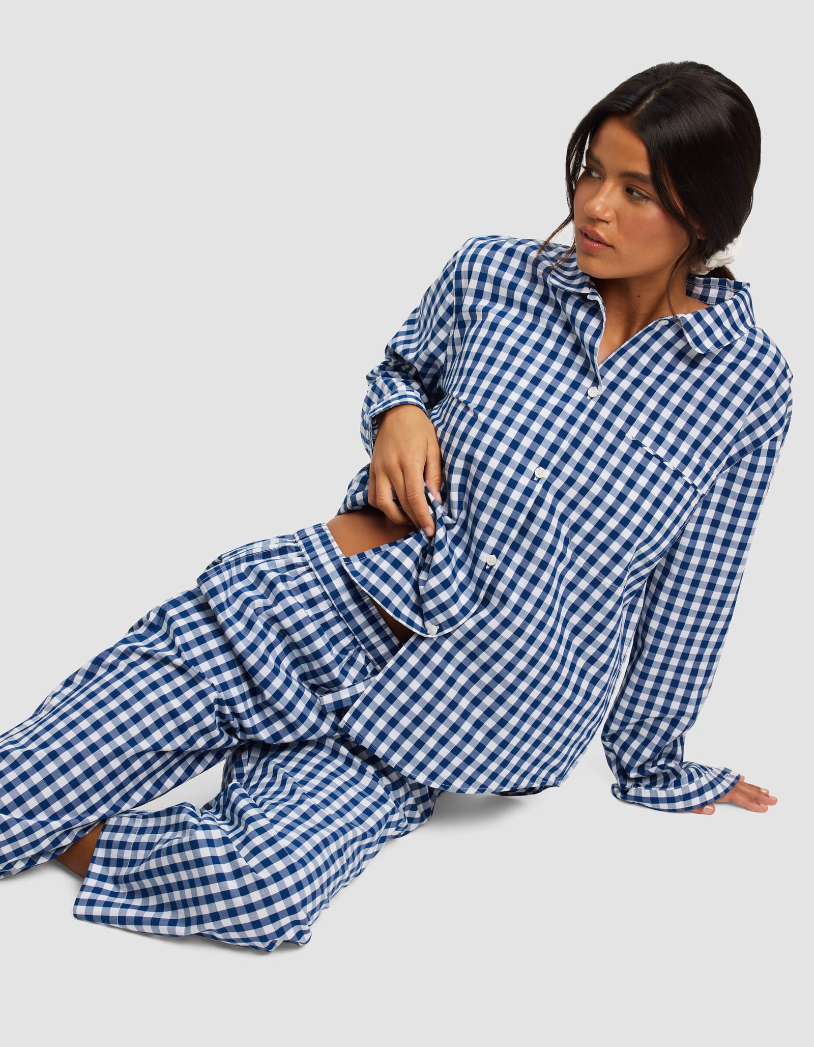 A woman with long dark hair wears the Cozy Earth Women's Sutton Long Sleeve Pajama Top in blue and white gingham, sitting on the floor and leaning to one side, looking off to her left against a plain, light background.