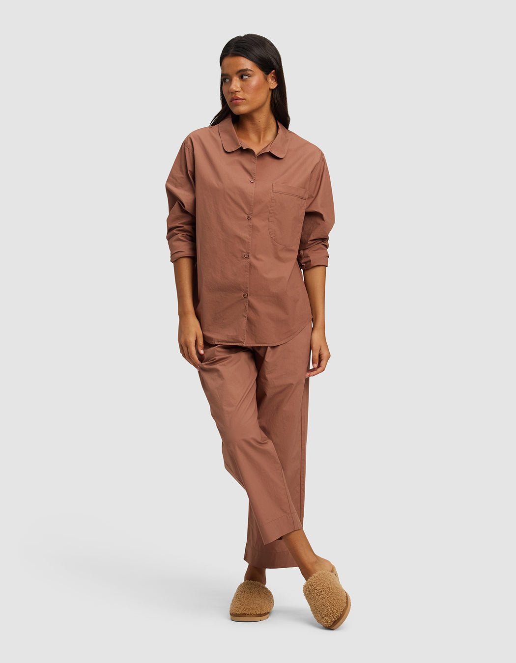 A woman with long dark hair and a neutral expression wears the Cozy Earth Women's Sutton Long Sleeve Pajama Top, styled against a plain backdrop with matching pants and beige slippers, one leg slightly crossed in front of the other.