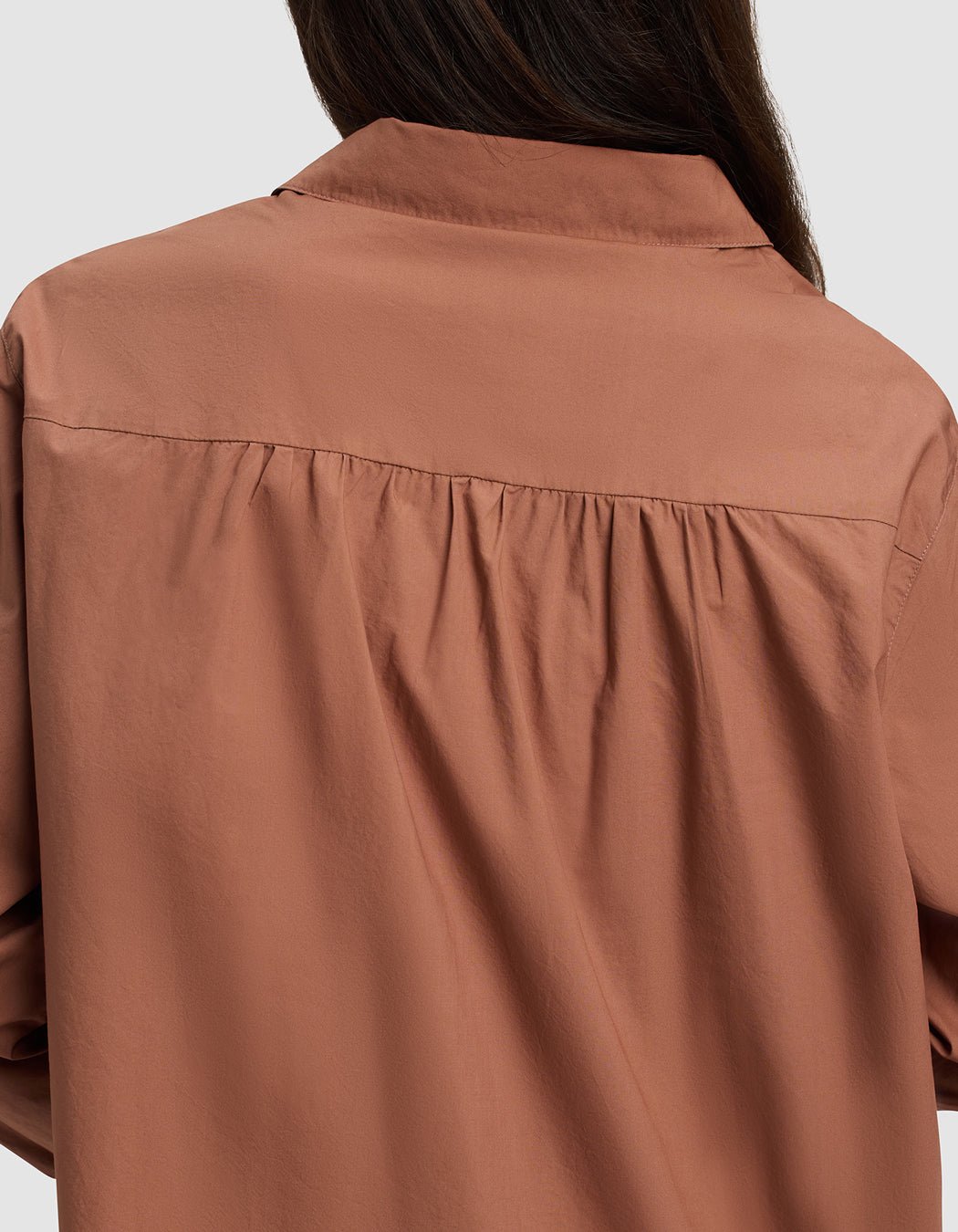 A woman with long brown hair is seen from behind wearing the Cozy Earth Women's Sutton Long Sleeve Pajama Top in rust, featuring loose fit and gathered fabric detail below the yoke.