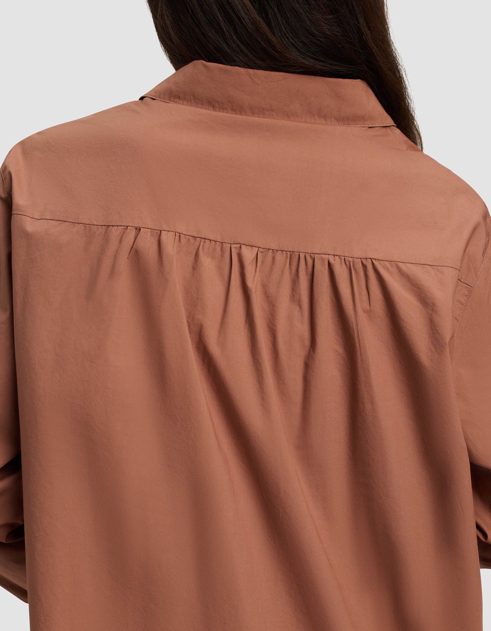 A close-up of the back of a person wearing the Cozy Earth Women's Sutton Long Sleeve Pajama Top, a loose brown shirt with gathered fabric below the shoulders. The person's dark hair shows against a plain light gray background.