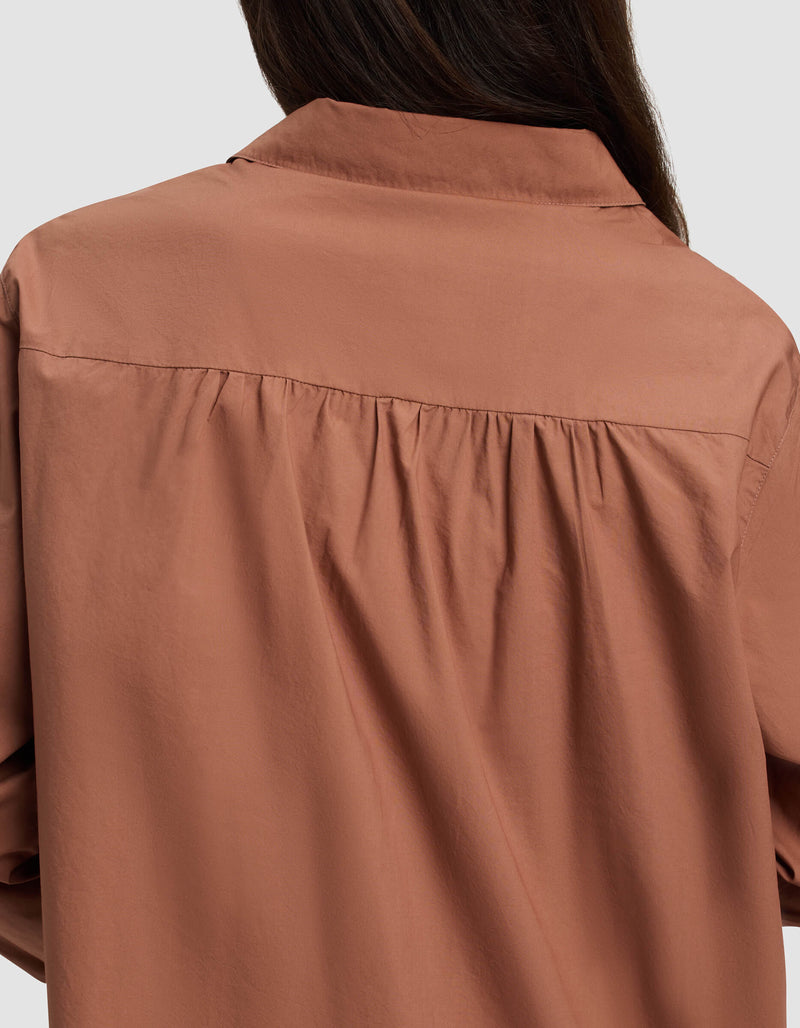 A close-up of the back of a person wearing the Cozy Earth Women's Sutton Long Sleeve Pajama Top, a loose brown shirt with gathered fabric below the shoulders. The person's dark hair shows against a plain light gray background.