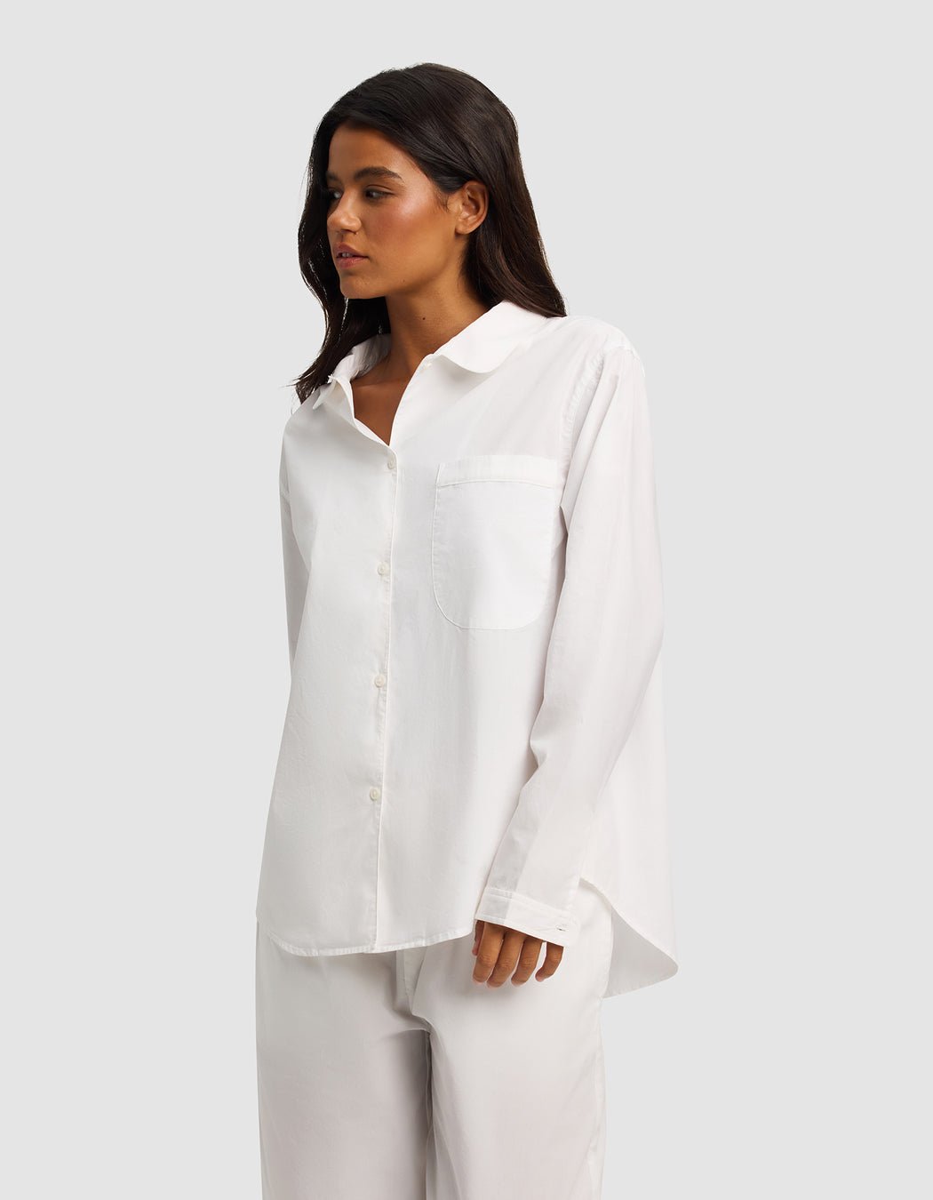 A woman with long dark hair wears the Cozy Earth Women's Sutton Long Sleeve Pajama Top— a loose-fitting white button-up with a chest pocket—paired with matching white pants, standing against a plain light gray background.
