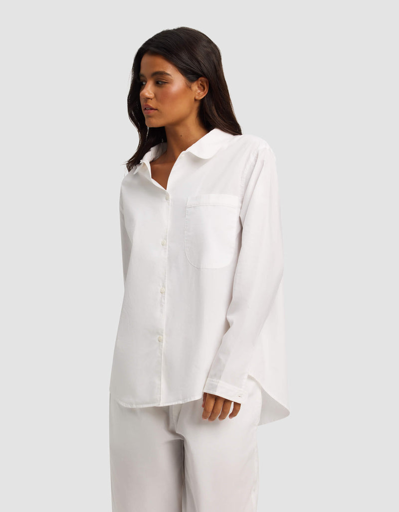 A woman with long dark hair wears the Cozy Earth Women's Sutton Long Sleeve Pajama Top, a loose-fitting white button-up with a chest pocket, paired with matching white pants, against a plain light gray background.