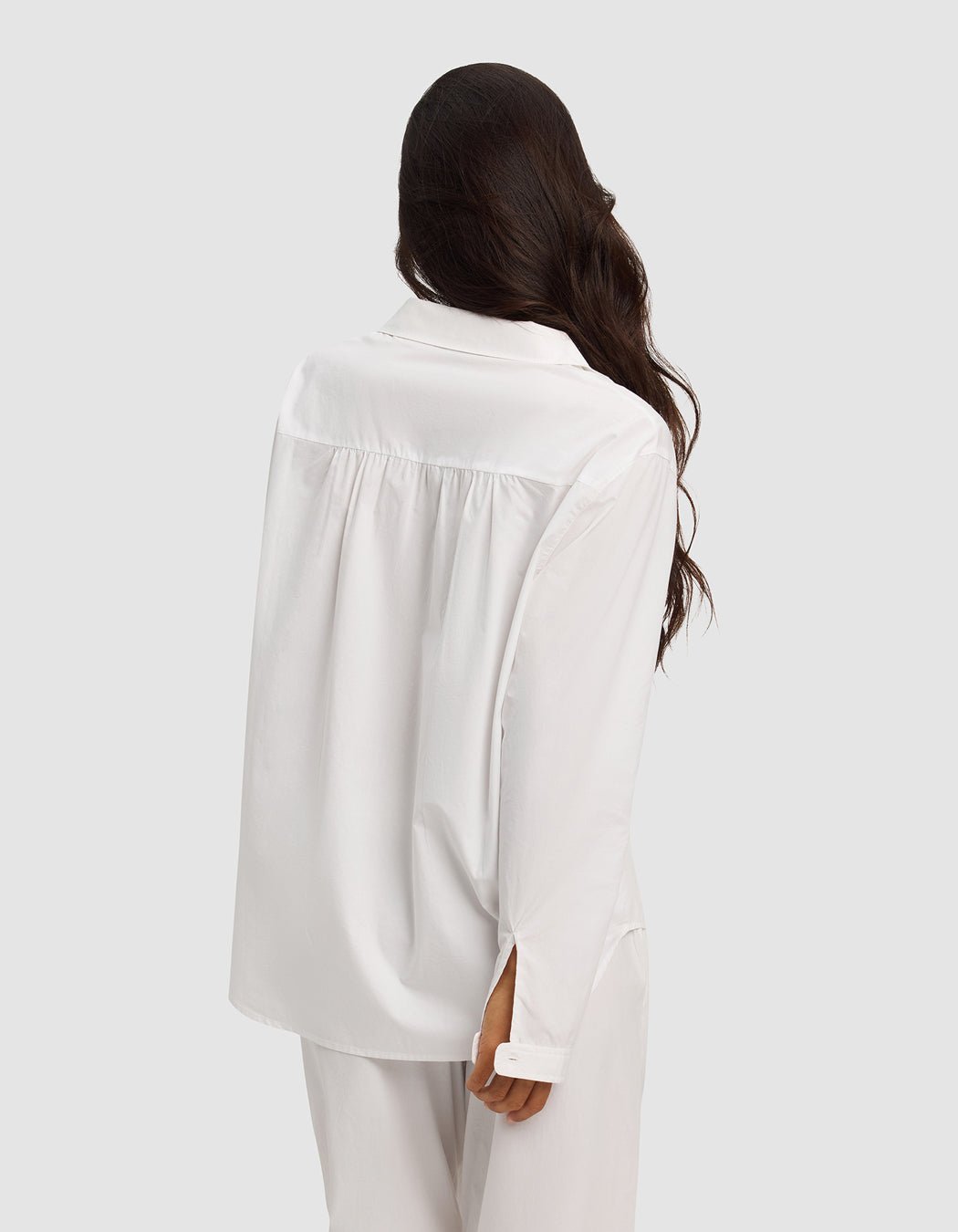 A woman with long wavy hair stands with her back to the camera, wearing the Cozy Earth Women's Sutton Long Sleeve Pajama Top and matching white pants against a plain light gray background.