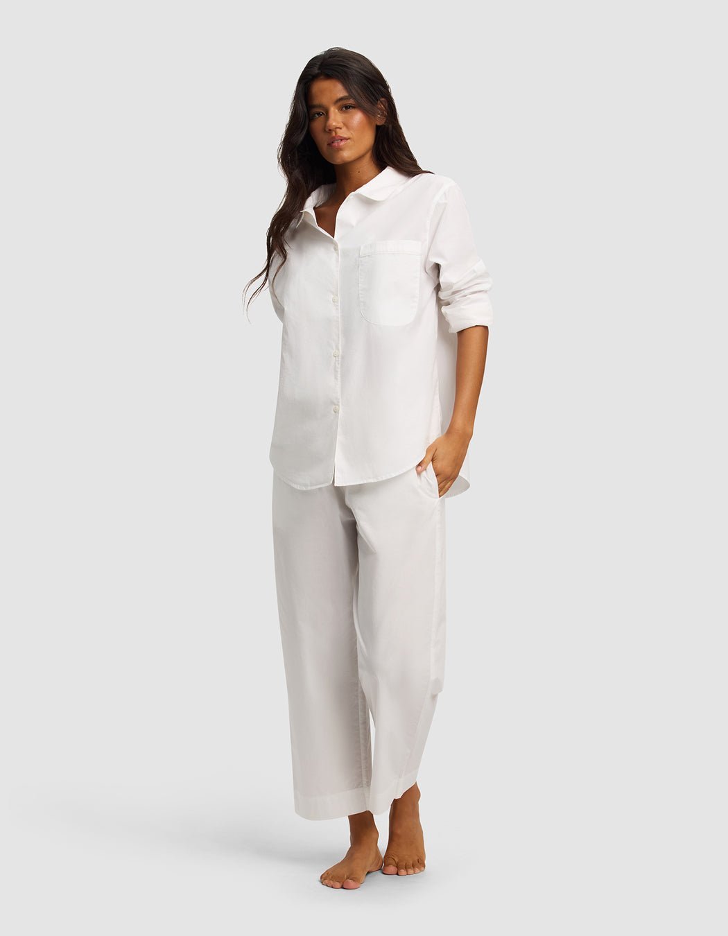 A woman with long dark hair stands barefoot against a plain background, wearing the Cozy Earth Women's Sutton Long Sleeve Pajama Top, a loose white button-up shirt with rolled sleeves, paired with matching wide-leg pants for a relaxed look.