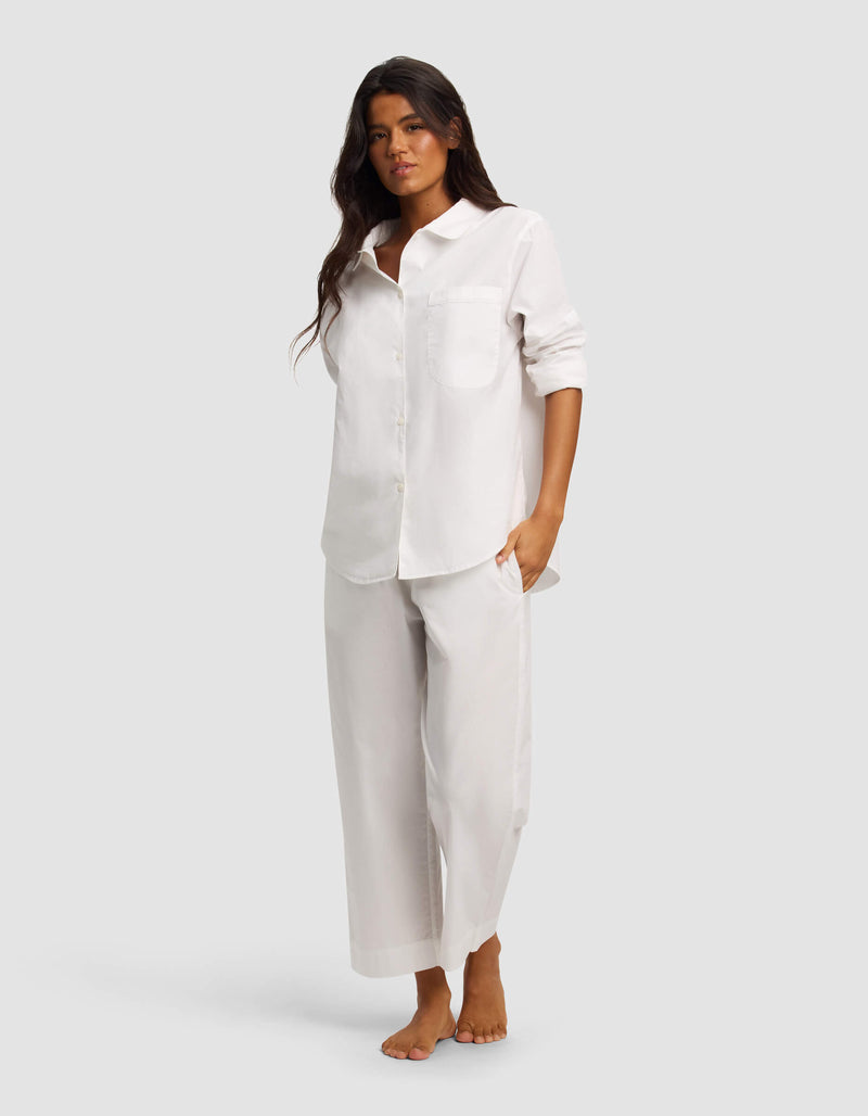 A woman with long dark hair stands barefoot against a plain background, wearing the Cozy Earth Women's Sutton Long Sleeve Pajama Top in white and matching wide-leg pants, styled with rolled sleeves and a relaxed, neutral expression.