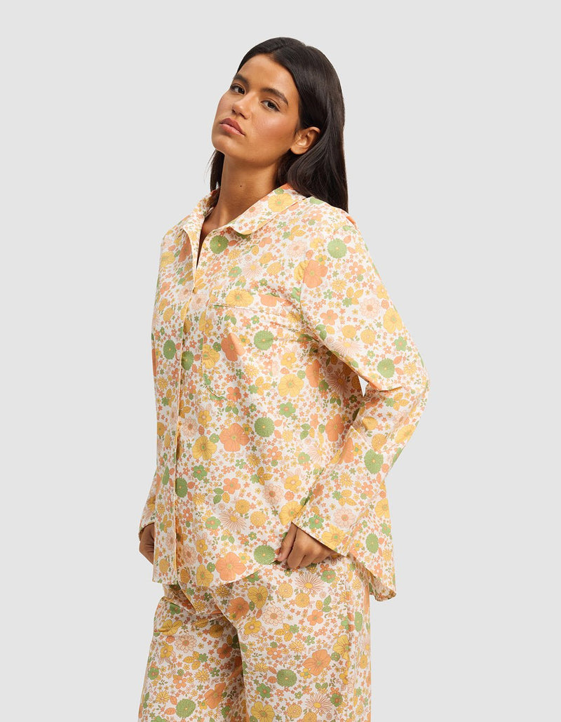 A woman with long dark hair models the Cozy Earth Women's Sutton Long Sleeve Pajama Top featuring a colorful floral print in orange, yellow, and green, standing with her hands in her pockets against a light gray backdrop.