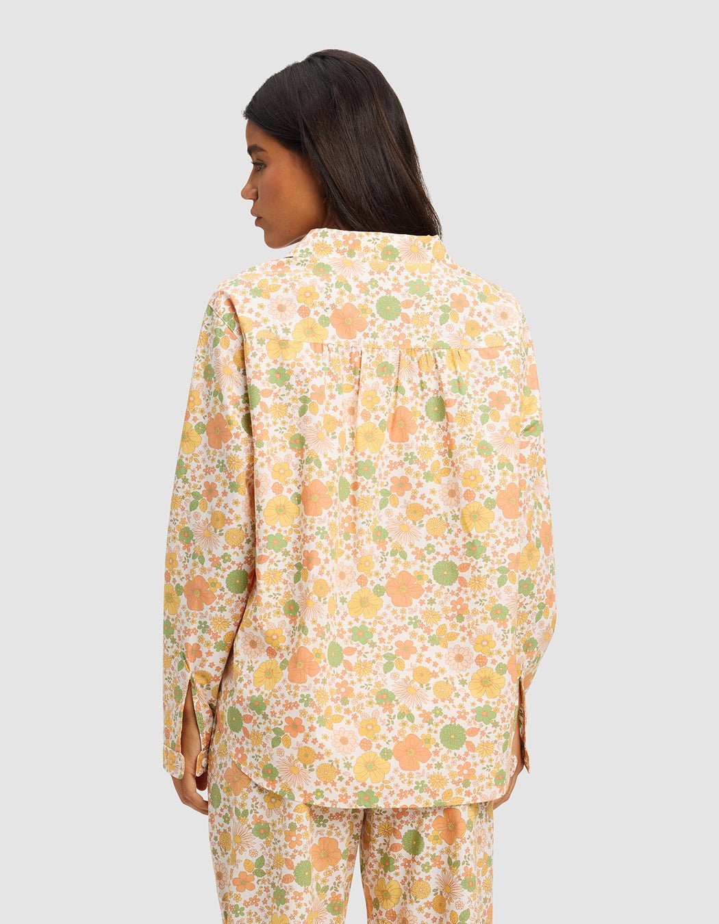 A person with long dark hair, seen from behind, wears the Cozy Earth Women's Sutton Long Sleeve Pajama Top in a loose floral pattern of pastel yellow, orange, and green. The background is plain light gray.