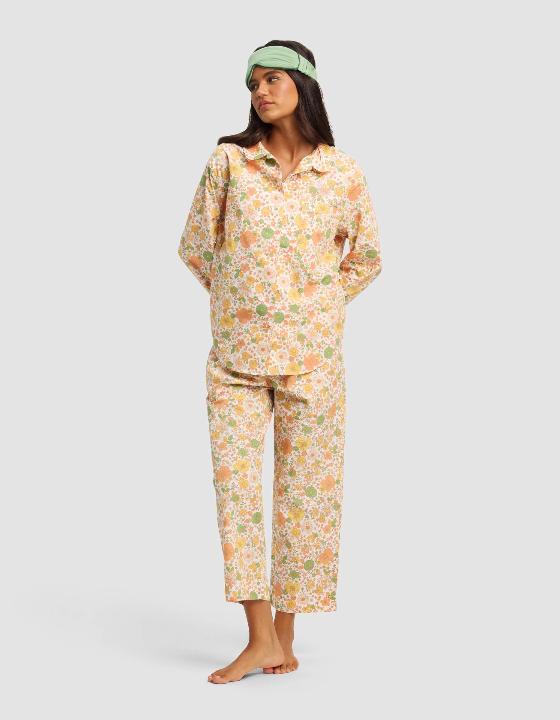 A woman stands barefoot against a plain light gray background, wearing the Cozy Earth Women’s Sutton Long Sleeve Pajama Top in a loose, floral pastel design and a green sleep mask.