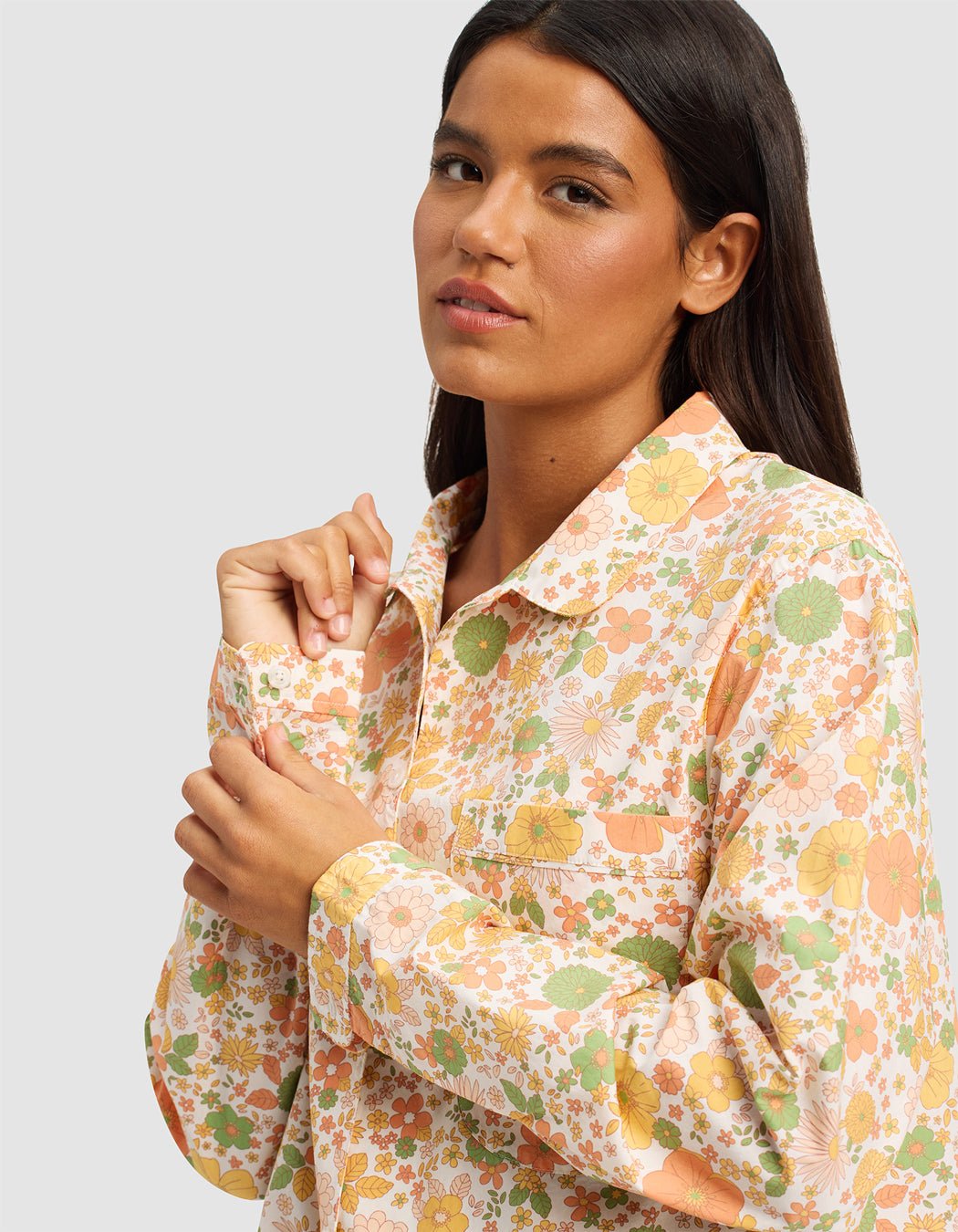 A woman with long dark hair wears the Women's Sutton Long Sleeve Pajama Top by Cozy Earth, featuring a colorful floral pattern in orange, yellow, and green, standing against a plain light gray background.