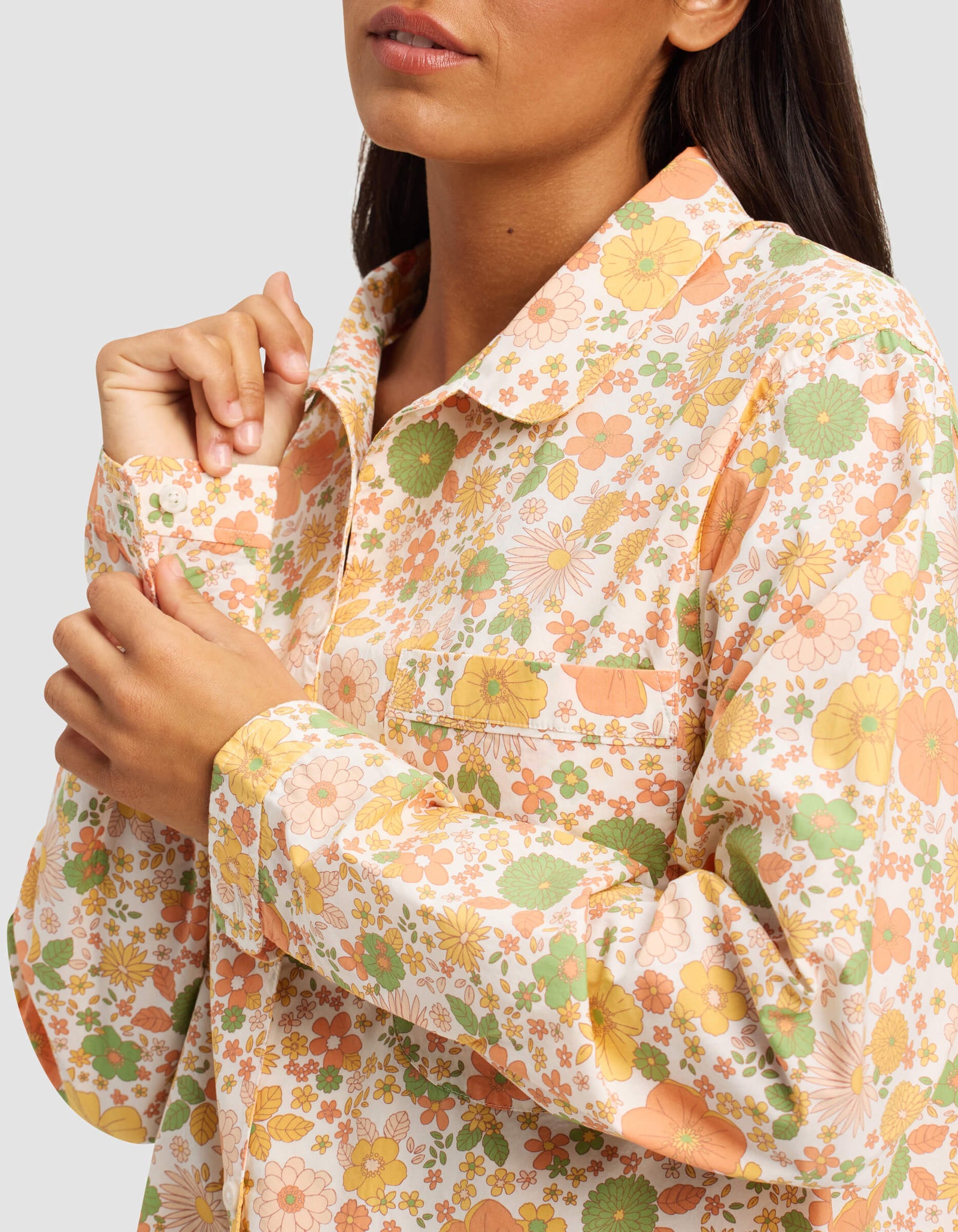 A woman buttons the collar of the Cozy Earth Women's Sutton Long Sleeve Pajama Top, featuring a colorful floral pattern in orange, green, and yellow. Her face is partially visible against a plain light gray background.