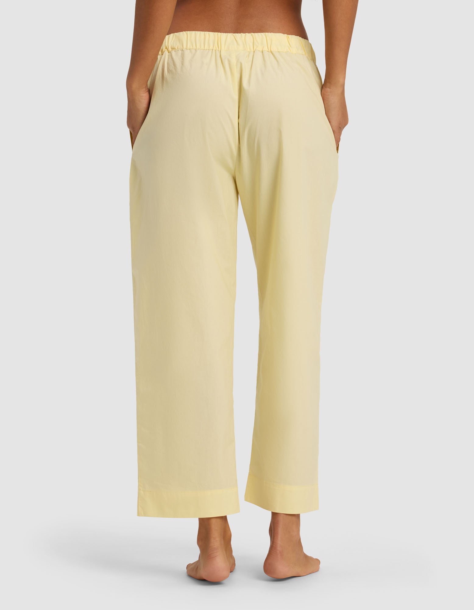 A person stands barefoot, facing away from the camera, wearing Cozy Earth's Women's Sutton Cropped Pajama Pant in light yellow with an elastic waistband and hands in the side pockets.