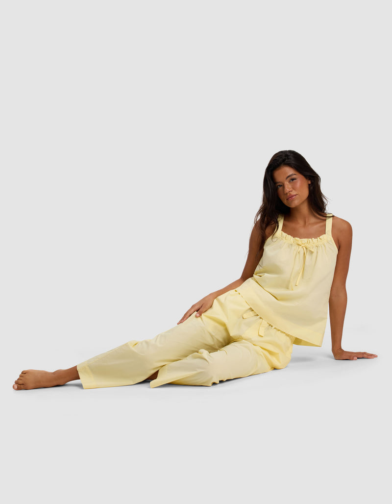 A woman with long dark hair sits on the floor against a light grey background, wearing Cozy Earth’s Women's Sutton Cropped Pajama Pant—a sleeveless yellow pajama set with loose-fitting pants and a ruffled neckline.