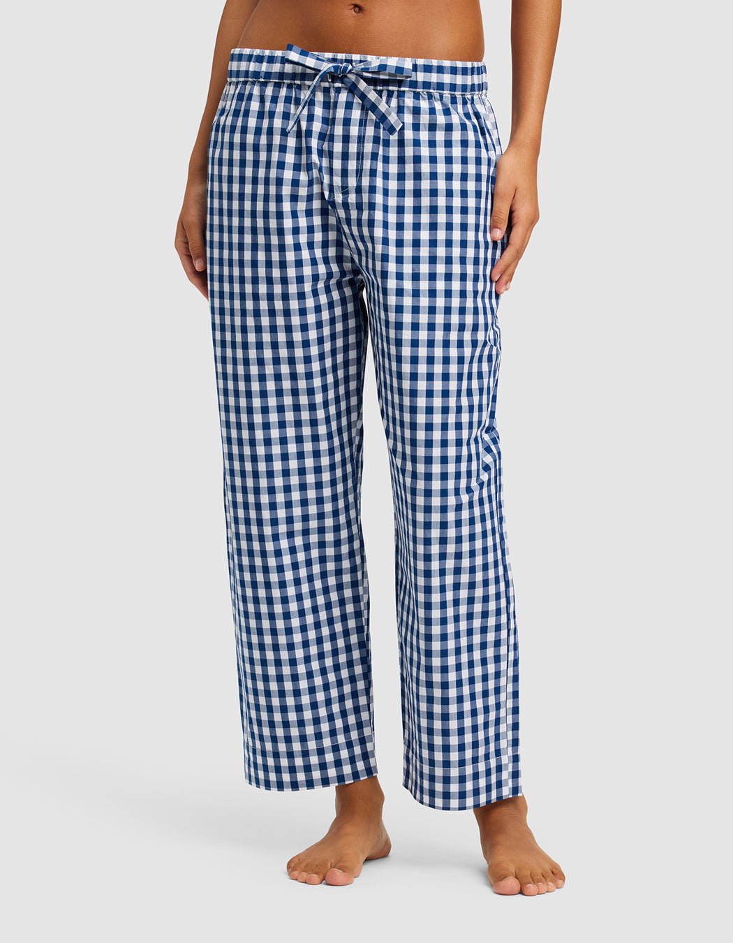 A woman wears Cozy Earth’s Women’s Sutton Cropped Pajama Pant, featuring a blue and white checkered pattern and drawstring waist, standing barefoot against a plain white background. 