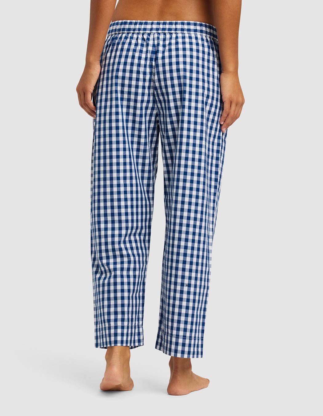 A woman stands barefoot, facing away, wearing Cozy Earth Women's Sutton Cropped Pajama Pant in blue and white gingham with an elastic waistband, set against a plain light background.