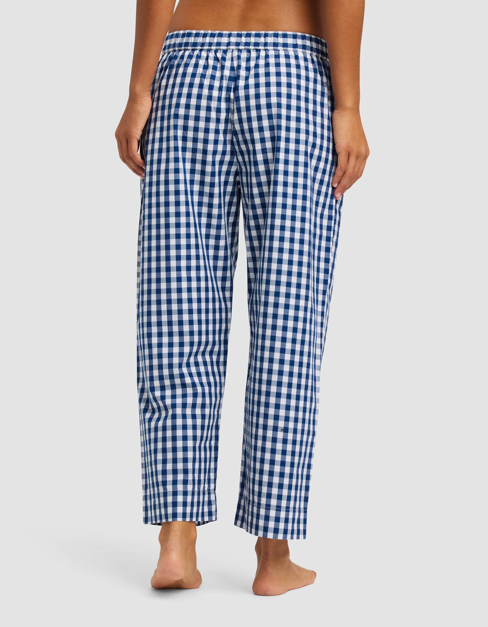 A woman stands barefoot, seen from behind, wearing Cozy Earth’s Women's Sutton Cropped Pajama Pant in blue and white check with an elastic waistband.