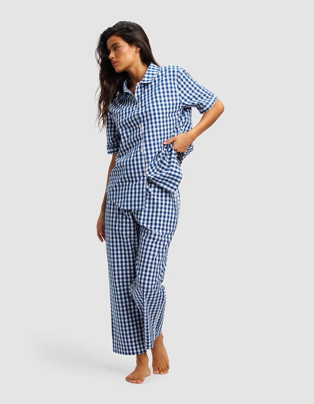 A woman stands barefoot against a plain light gray background, wearing Cozy Earth's Women's Sutton Cropped Pajama Pant—a blue and white checkered set with short sleeves and cropped pants.