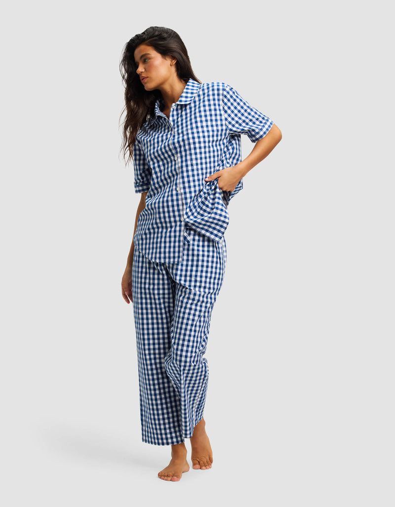 A woman stands barefoot in Cozy Earth’s Women's Sutton Cropped Pajama Pant set, featuring a blue and white checkered short-sleeved button-up shirt with loose pants, against a plain light gray background.