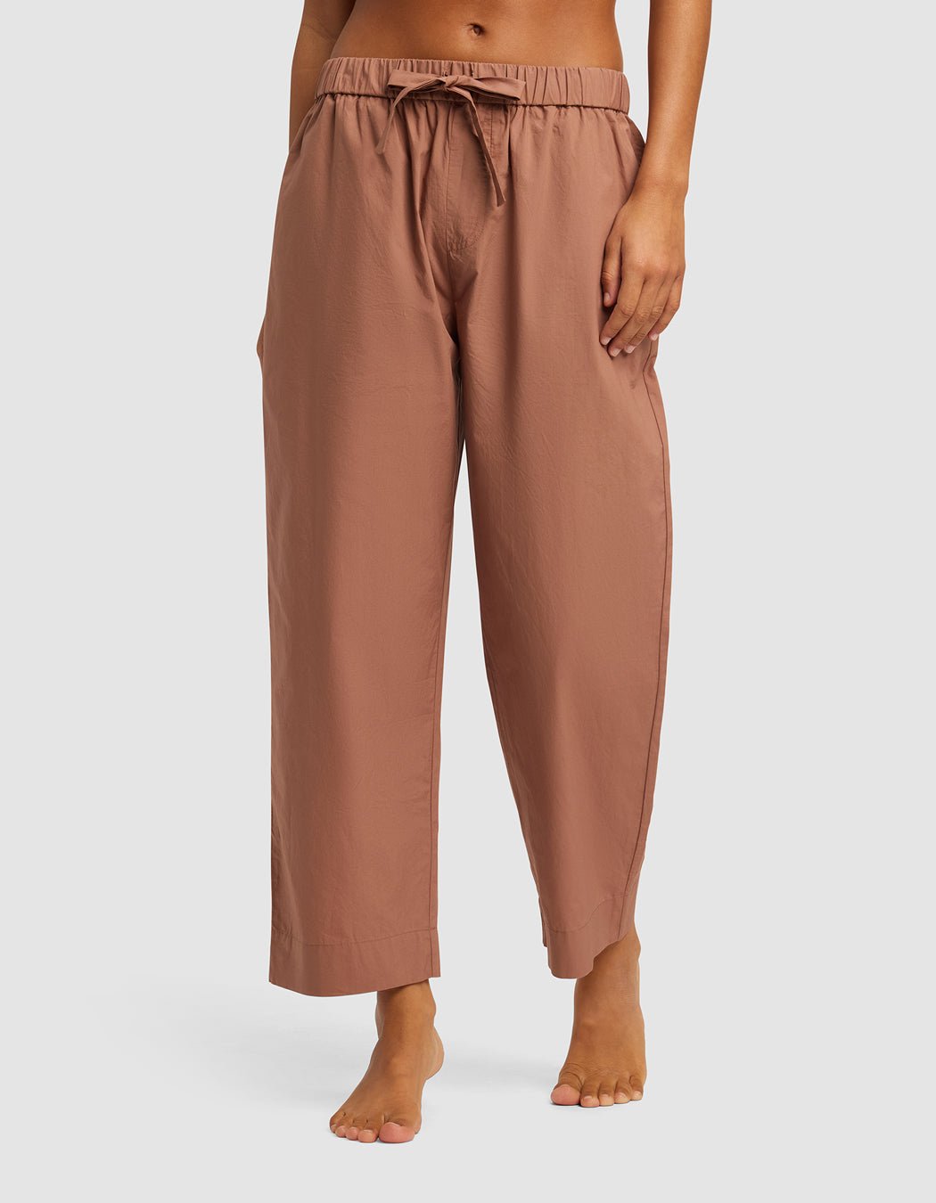 A woman models Cozy Earth's Women's Sutton Cropped Pajama Pant—loose, light brown, ankle-length with an elastic waistband and drawstring—while standing barefoot against a plain background.|Color:Saddle