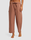 A woman stands barefoot in Cozy Earth’s Women’s Sutton Cropped Pajama Pant—loose, light brown drawstring pants with side pockets, cropped above the ankles—against a plain light background.|Color:Saddle