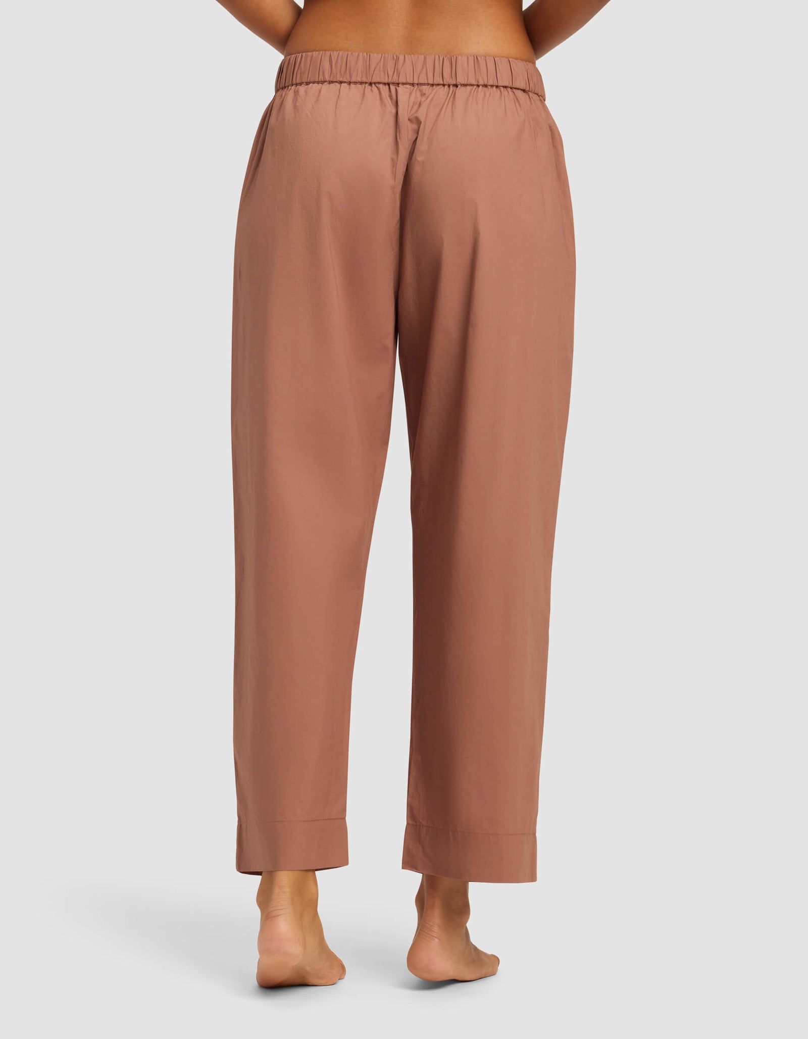 A person is barefoot on a light surface, shown from the back, wearing Cozy Earth’s Women's Sutton Cropped Pajama Pant—loose-fitting, high-waisted pajama pants in light brown or terracotta with an elastic waistband and relaxed straight legs.
